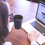 Administrativo 100% Remoto e + vagas abertas na FCamara A woman enjoys coffee while browsing the web on a laptop at a home office desk.