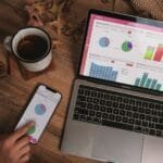 Assistente de Customer Success com VR/VA até R$ 1.500 e +Remotas A laptop and smartphone displaying analytics dashboards on a wooden desk with a mug